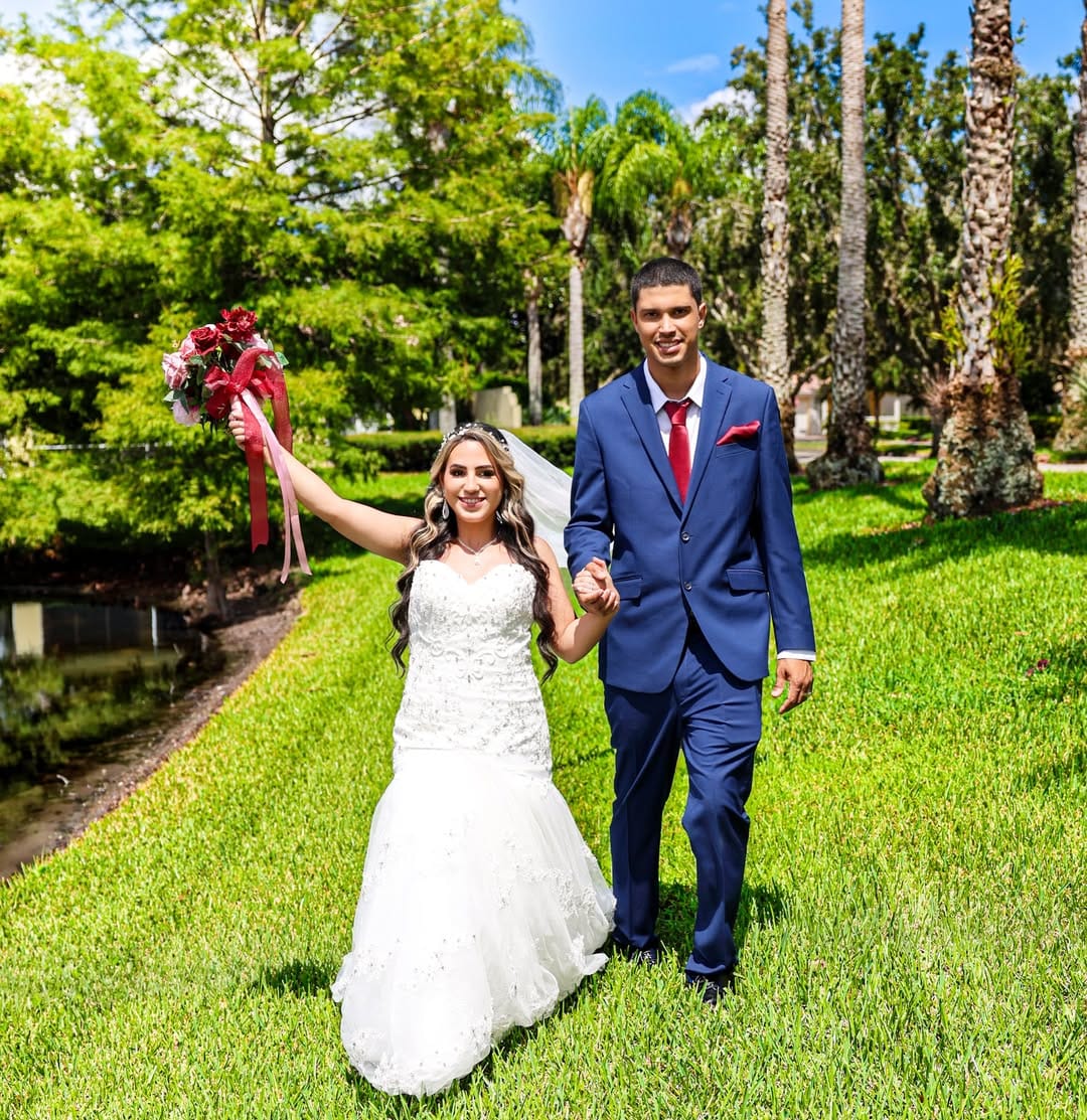 Bride and groom holding hands and smiling while walking across a sunny green lawn, with the bride raising a red floral bouquet and the groom wearing a blue suit with red accents.