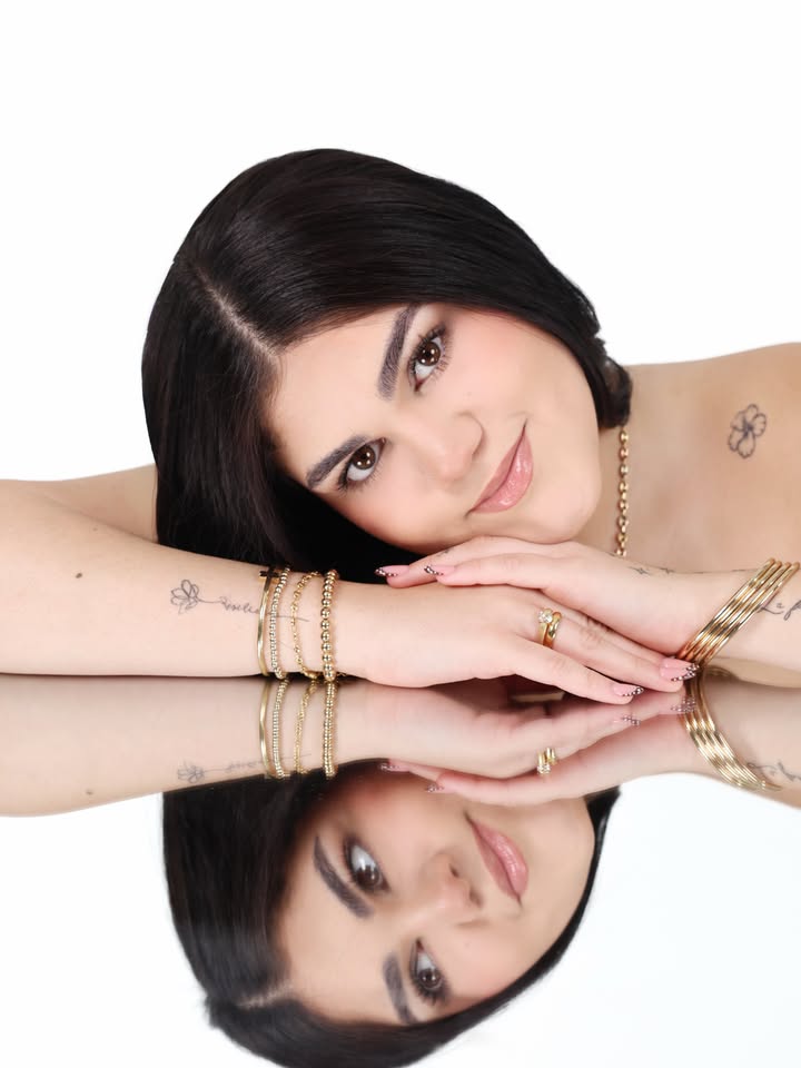 A woman rests her head on her folded arms over a reflective surface, creating a clear mirror-like reflection below. She has straight dark hair, subtle makeup, visible arm and shoulder tattoos, and wears several gold-toned bracelets, rings, and a necklace against a white background.
