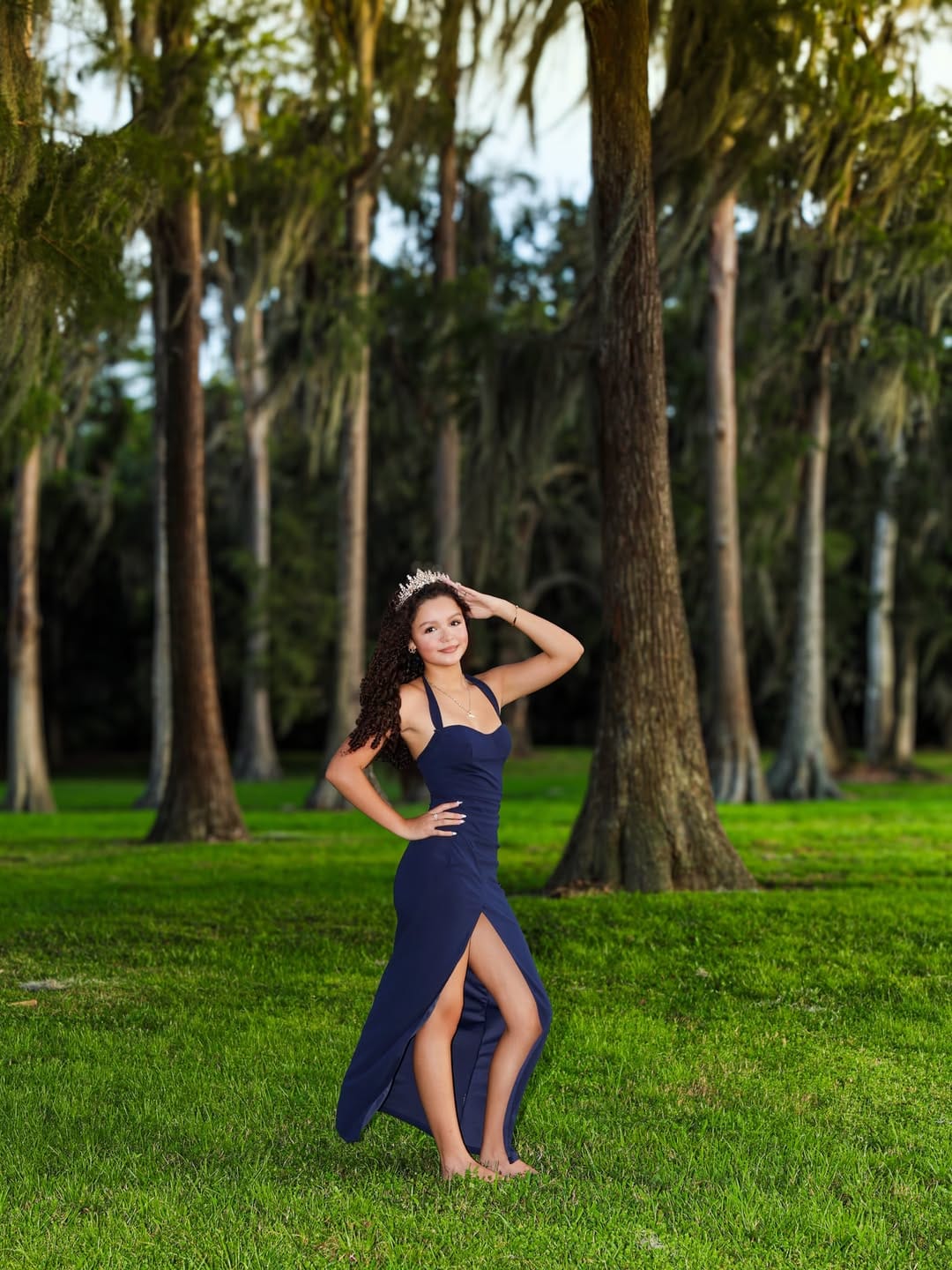 Young quinceañera wearing a navy blue gown and tiara, posing barefoot on a green lawn surrounded by tall trees in a sunlit park.
