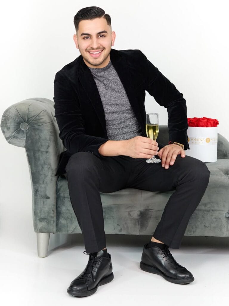 Smiling man in a black blazer and gray shirt seated on a gray velvet sofa, holding a glass of champagne beside a box of red roses against a white studio background.