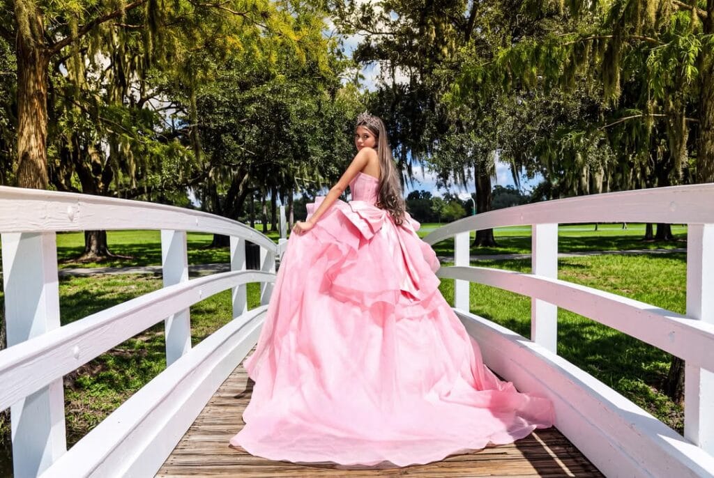 Young quinceañera in a voluminous pink ball gown and tiara standing on a white wooden bridge, looking back toward the camera in a sunny green park surrounded by large trees.